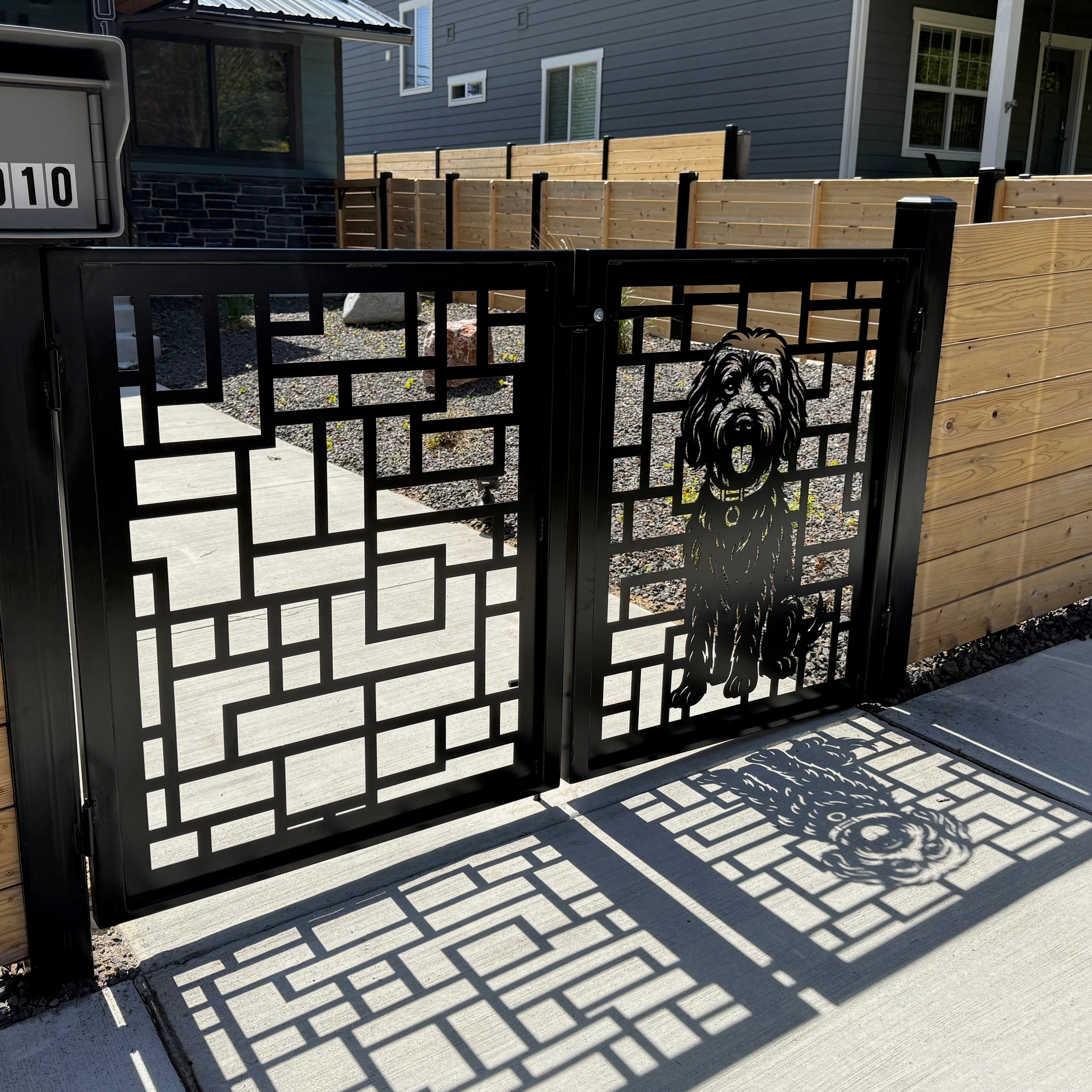 Decorative metal gate with geometric design in front of a wooden fence, house in the background
